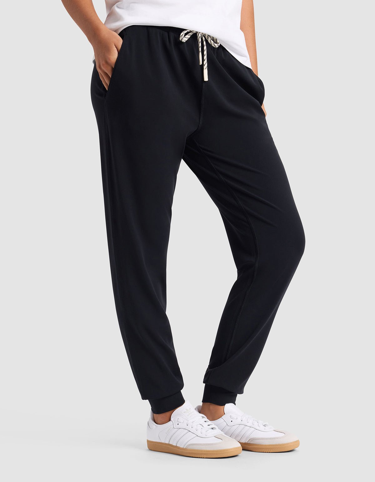 A woman wears Cozy Earth Women's Studio Jogger—black with a drawstring waist—paired with white sneakers and a white top, standing hands-in-pockets against a plain light gray background.