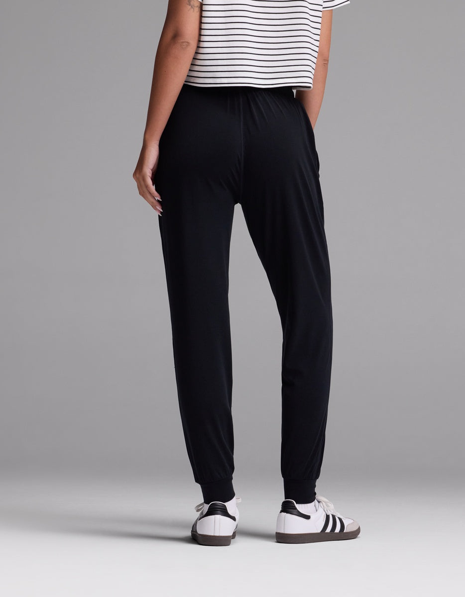 A woman is standing against a gray background, wearing Cozy Earth’s Women’s Studio Jogger pants in black, paired with a white and black striped short-sleeve shirt and white sneakers with black stripes. The photo is taken from the back.