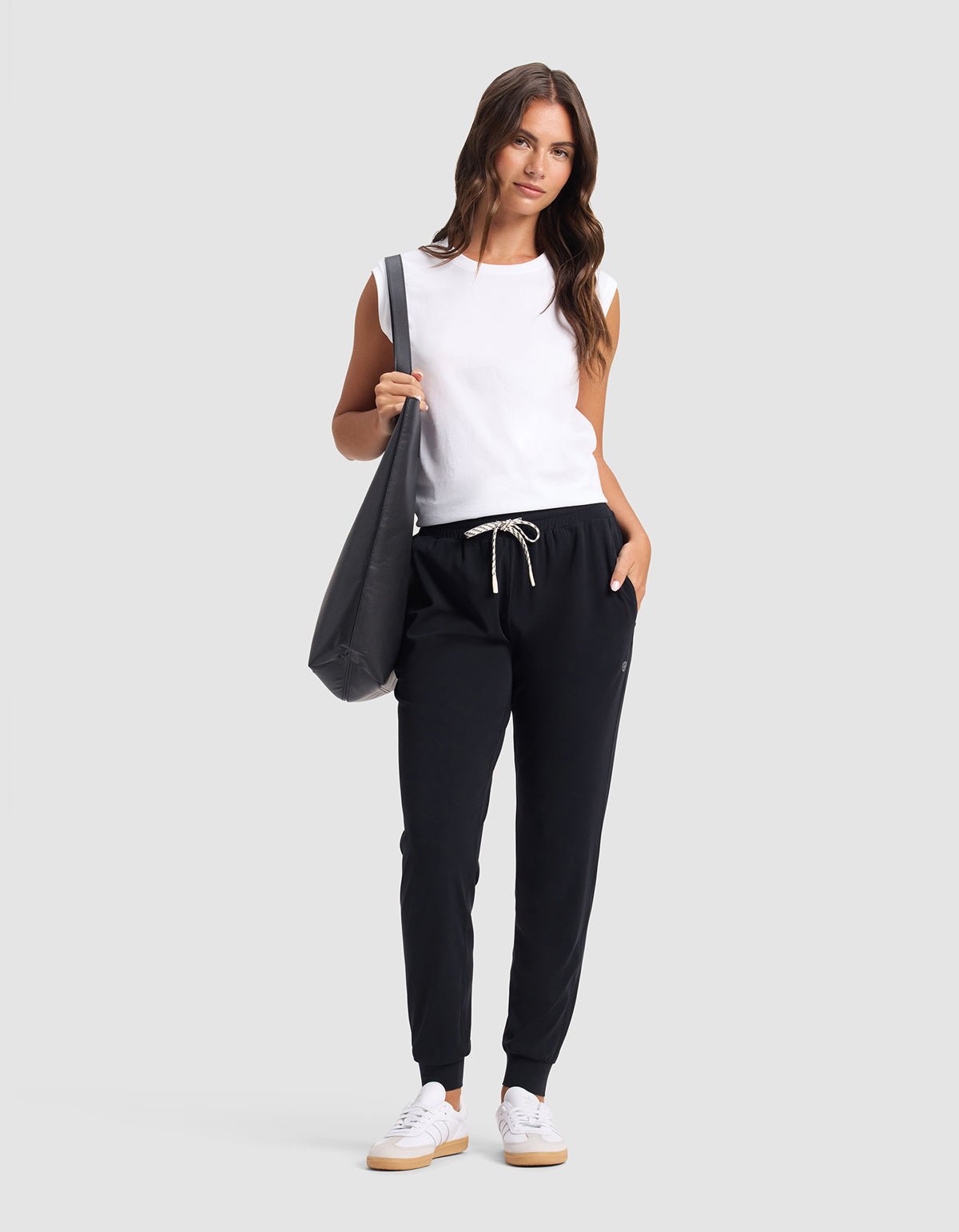 A woman poses against a plain background wearing Cozy Earth's Women's Studio Jogger in black with a drawstring, a sleeveless white top, white sneakers, and carrying a large black tote bag on her shoulder.
