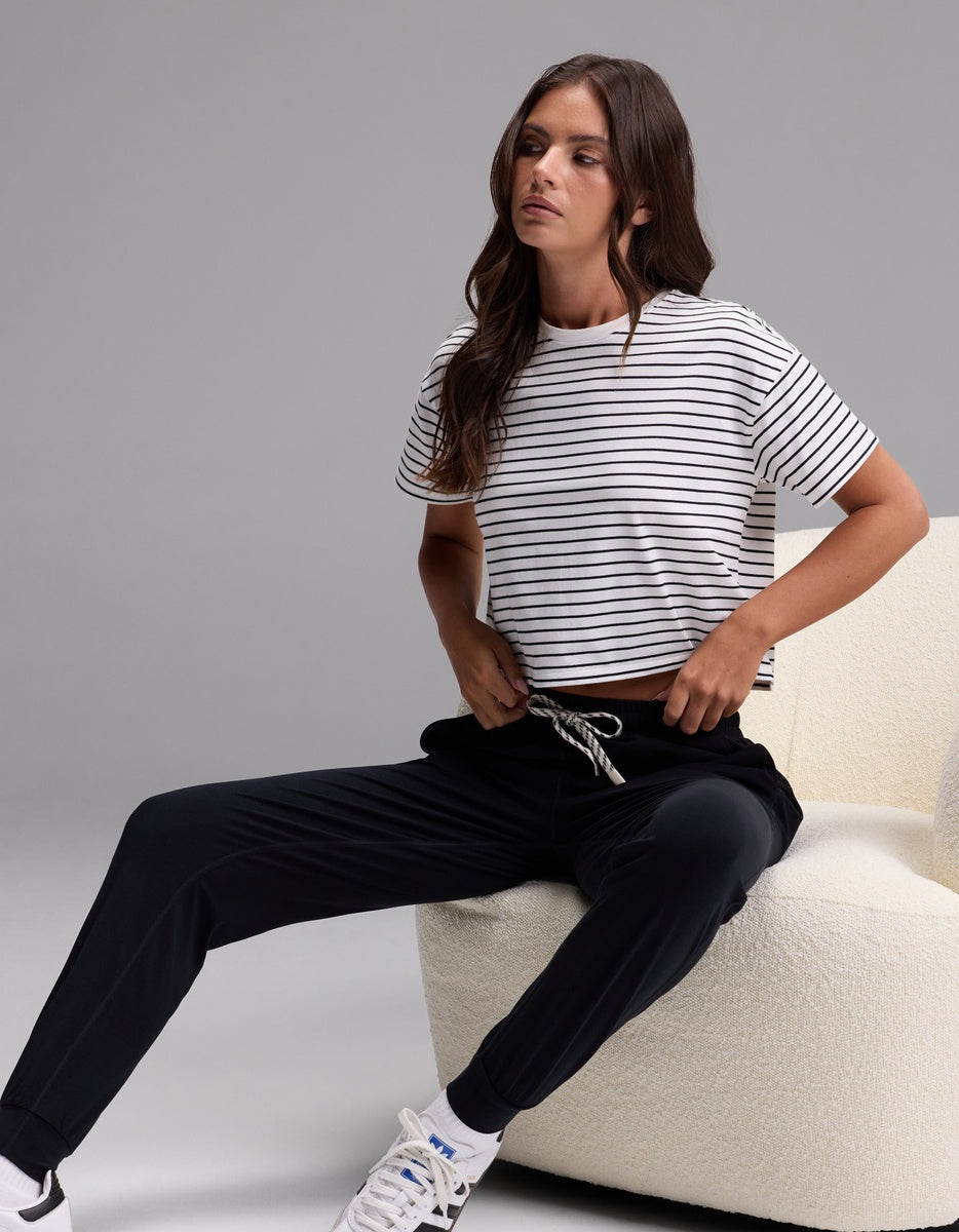 A woman with long brown hair and a relaxed expression sits on a white chair against a plain gray background, wearing Cozy Earth’s Women’s Studio Jogger in black, paired with a black and white striped t-shirt and white sneakers.