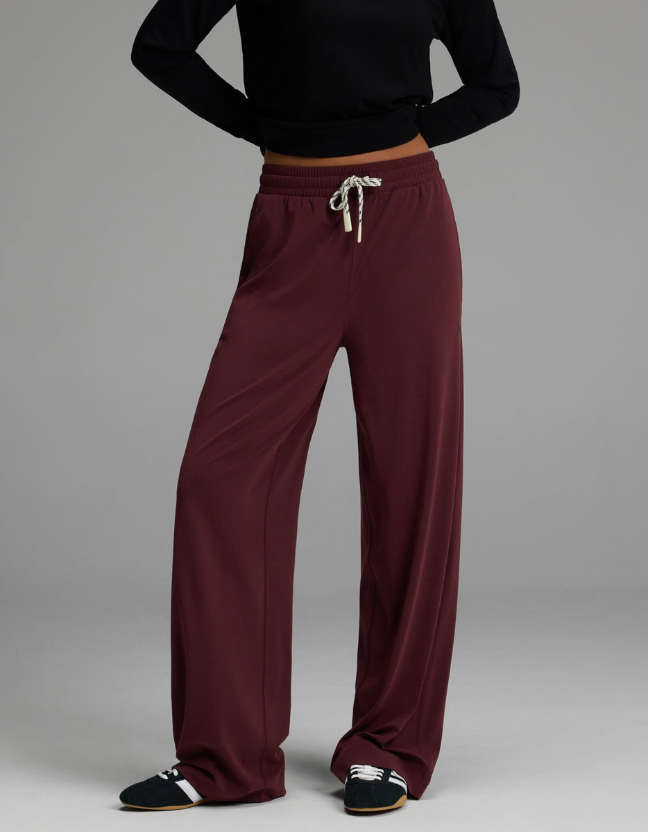 A person wearing a black long-sleeve top, Cozy Earth Women’s Studio Wide Leg Pant in burgundy, and black slip-on shoes with white and beige stripes stands against a plain gray background.