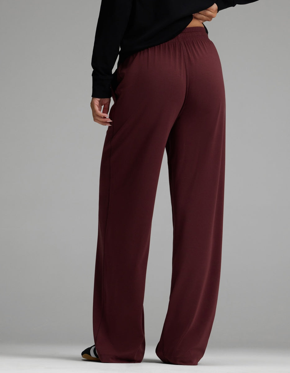 A person wearing Cozy Earth’s Women’s Studio Wide Leg Pant in maroon and a black long-sleeve top stands against a plain gray background, facing away from the camera.