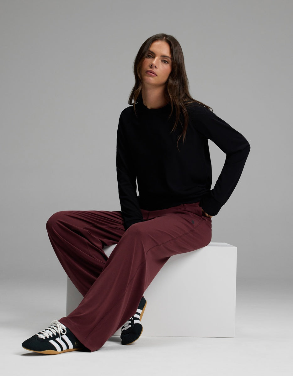 A person with long brown hair sits on a white block, wearing a black long-sleeve shirt, Cozy Earth’s Women’s Studio Wide Leg Pant in maroon, and black Adidas sneakers with white stripes, against a plain gray background.