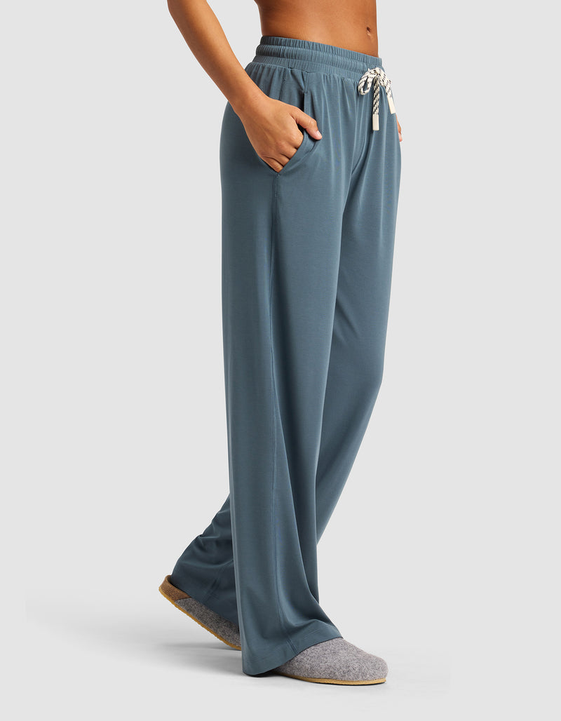 A woman stands with hands in the pockets of Cozy Earth's Women's Studio Wide Leg Pant—loose-fitting, blue drawstring lounge pants—paired with gray slippers against a plain, light background.