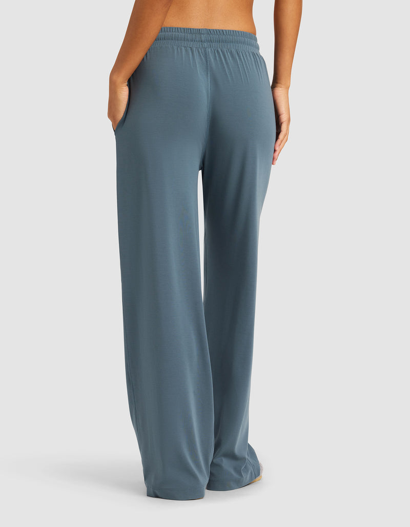 A person is shown from the back, barefoot against a plain background, wearing Cozy Earth’s Women's Studio Wide Leg Pant in loose-fitting blue-gray with an elastic waistband and side pockets.