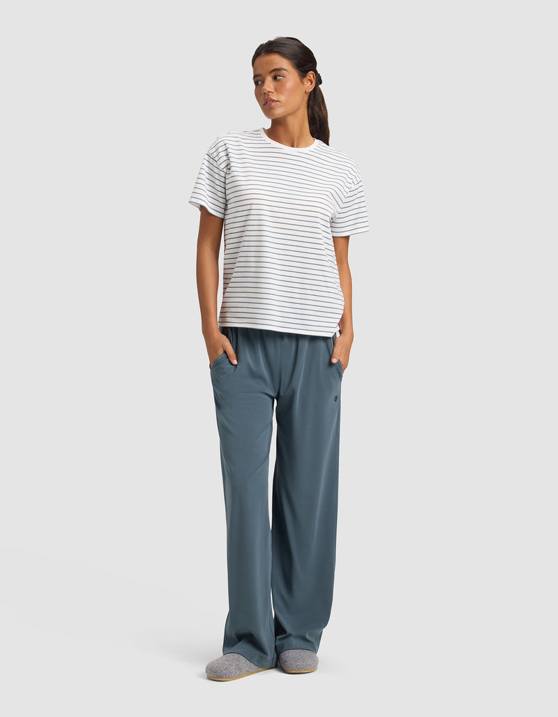 A woman stands with hands in her pockets, wearing Cozy Earth's Women's Studio Wide Leg Pant in blue, paired with a white and gray striped short-sleeve t-shirt and gray slippers against a plain light background.