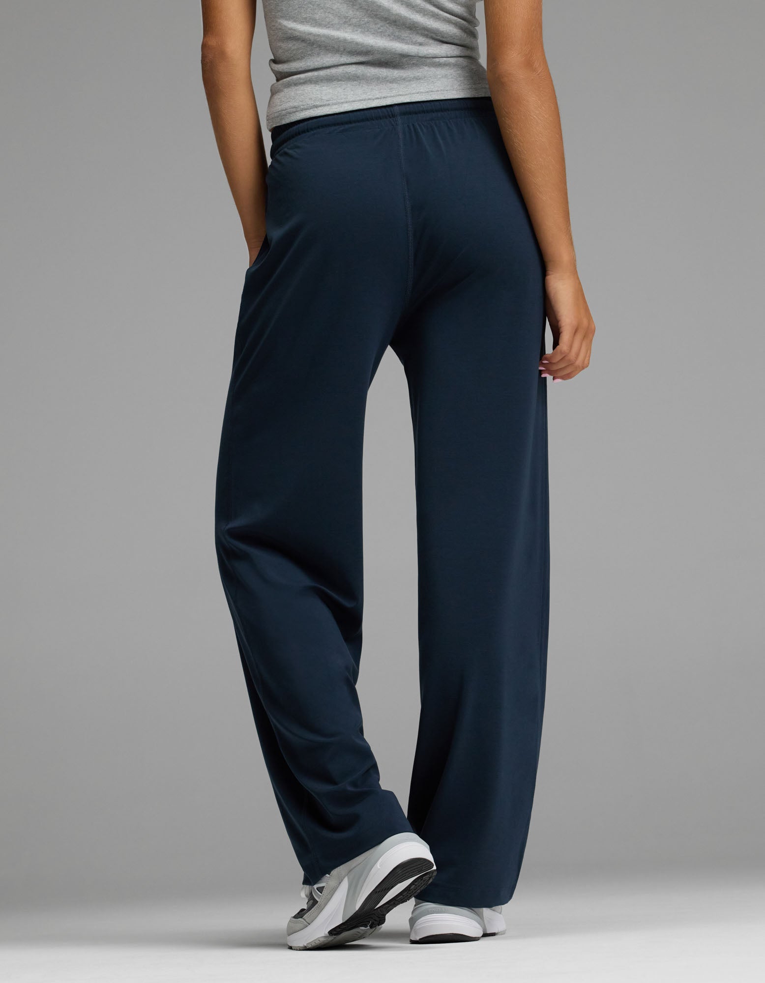 A person is seen from behind, standing against a plain grey background with one hand in their pocket, wearing the Cozy Earth Women’s Studio Wide Leg Pant in navy blue, paired with a grey top and white sneakers.