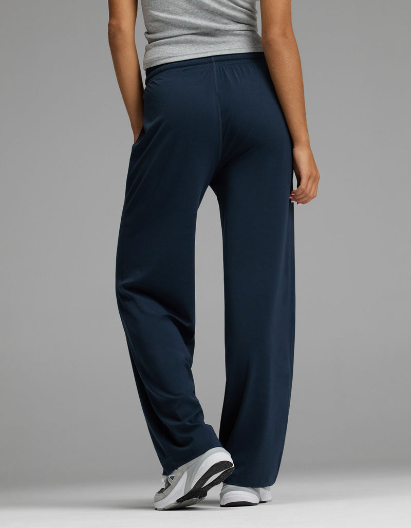 A person is seen from behind, standing against a plain grey background with one hand in their pocket, wearing the Cozy Earth Women’s Studio Wide Leg Pant in navy blue, paired with a grey top and white sneakers.