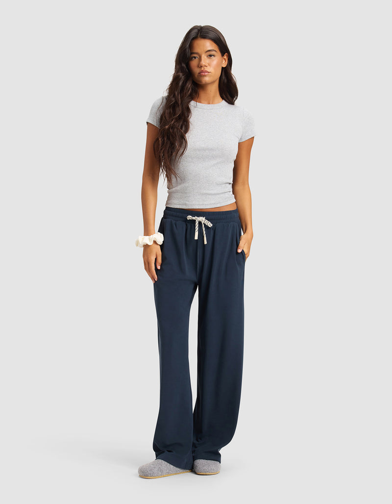 A woman with long dark hair wears Cozy Earth's Women's Studio Wide Leg Pant in navy blue, a fitted gray short-sleeve top, gray slippers, and a scrunchie on her wrist. She stands against a plain light gray background.