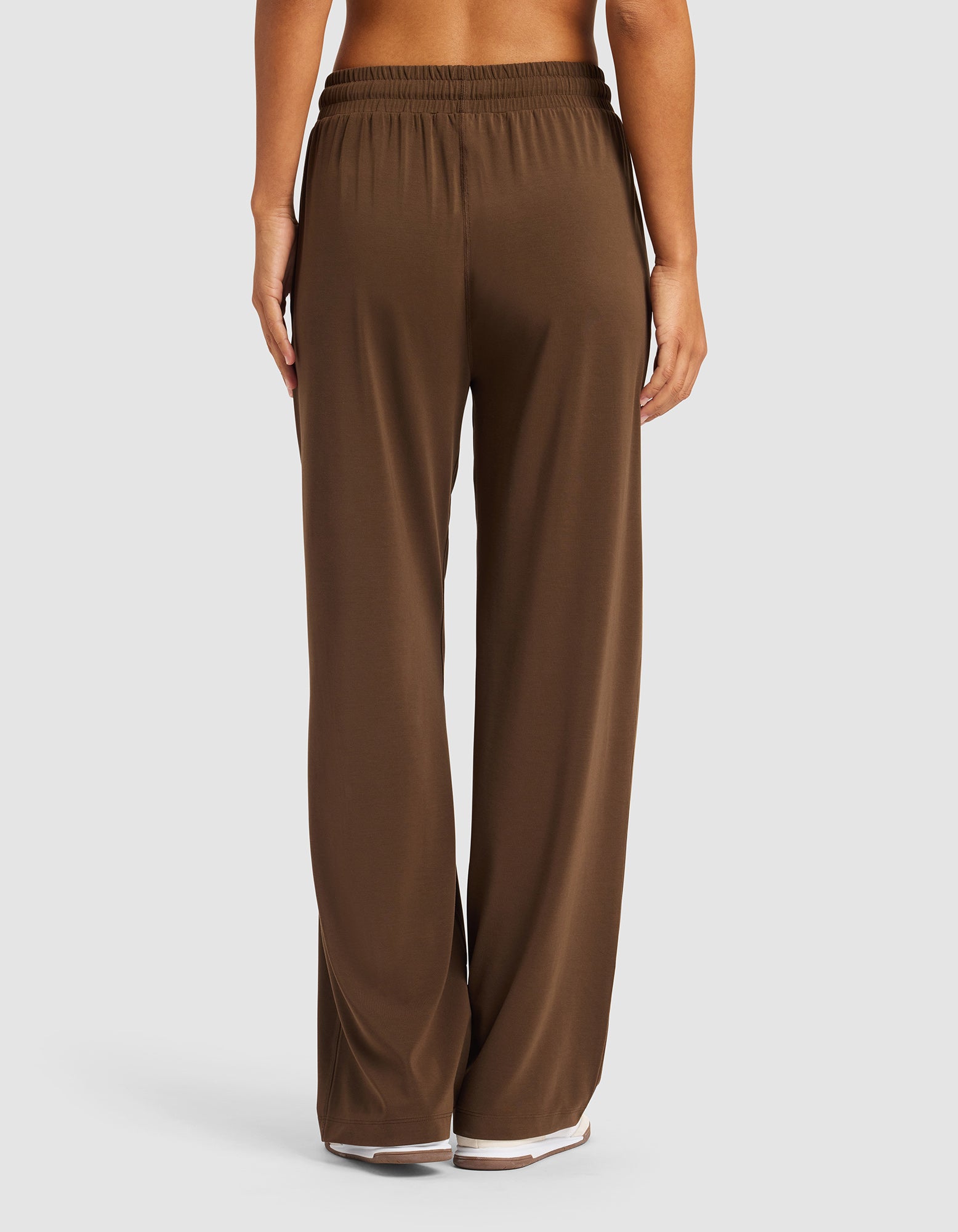 A woman is shown from the back, barefoot on a white background, wearing Cozy Earth’s Women's Studio Wide Leg Pant in brown with a loose fit and elastic waistband.