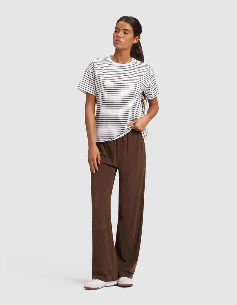 A woman stands against a plain background wearing Cozy Earth Women's Studio Wide Leg Pants in brown, a white and black striped short-sleeve t-shirt, and white slip-on shoes. Her long dark hair is pulled back, and her left hand is in her pocket.
