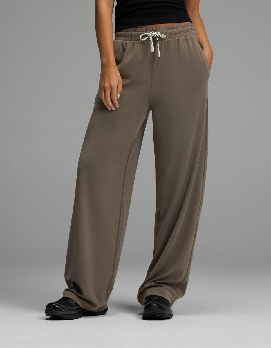 A person wearing Cozy Earth’s Women’s Studio Wide Leg Pant in Granite, paired with a black top and black shoes, stands with hands in pockets against a plain gray background.