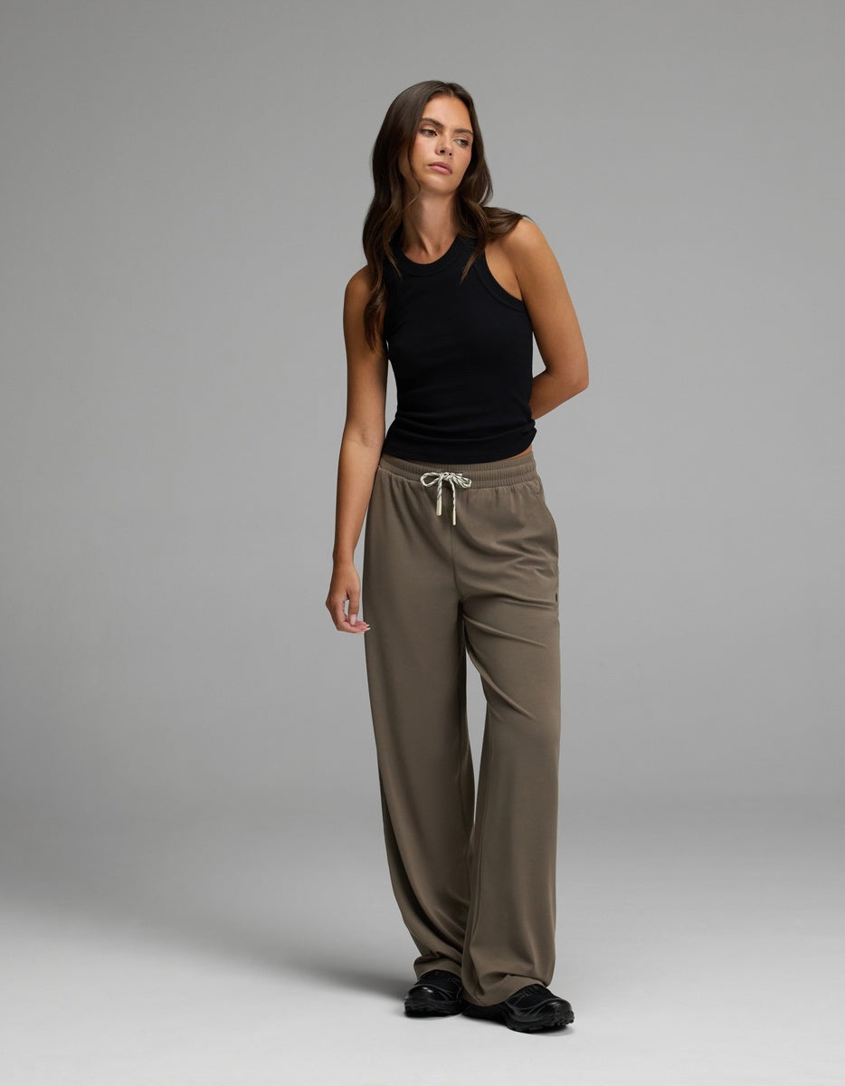 A woman stands in a studio against a plain gray background, wearing Cozy Earth’s Women’s Studio Wide Leg Pant in taupe, paired with a black sleeveless top and black shoes.