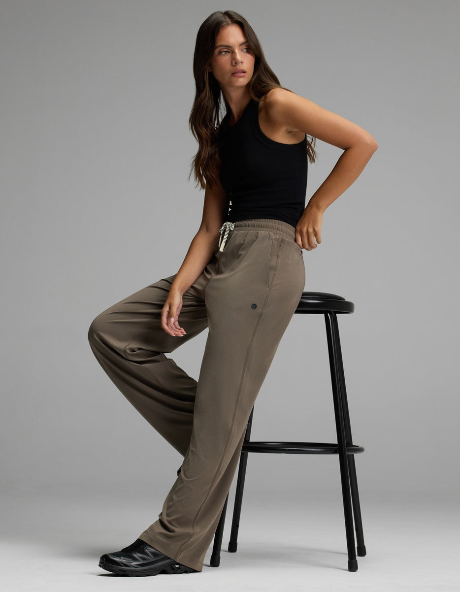 A woman models the Cozy Earth Women’s Studio Wide Leg Pant in taupe, paired with a black sleeveless top as she sits confidently on a tall black stool against a plain grey background.