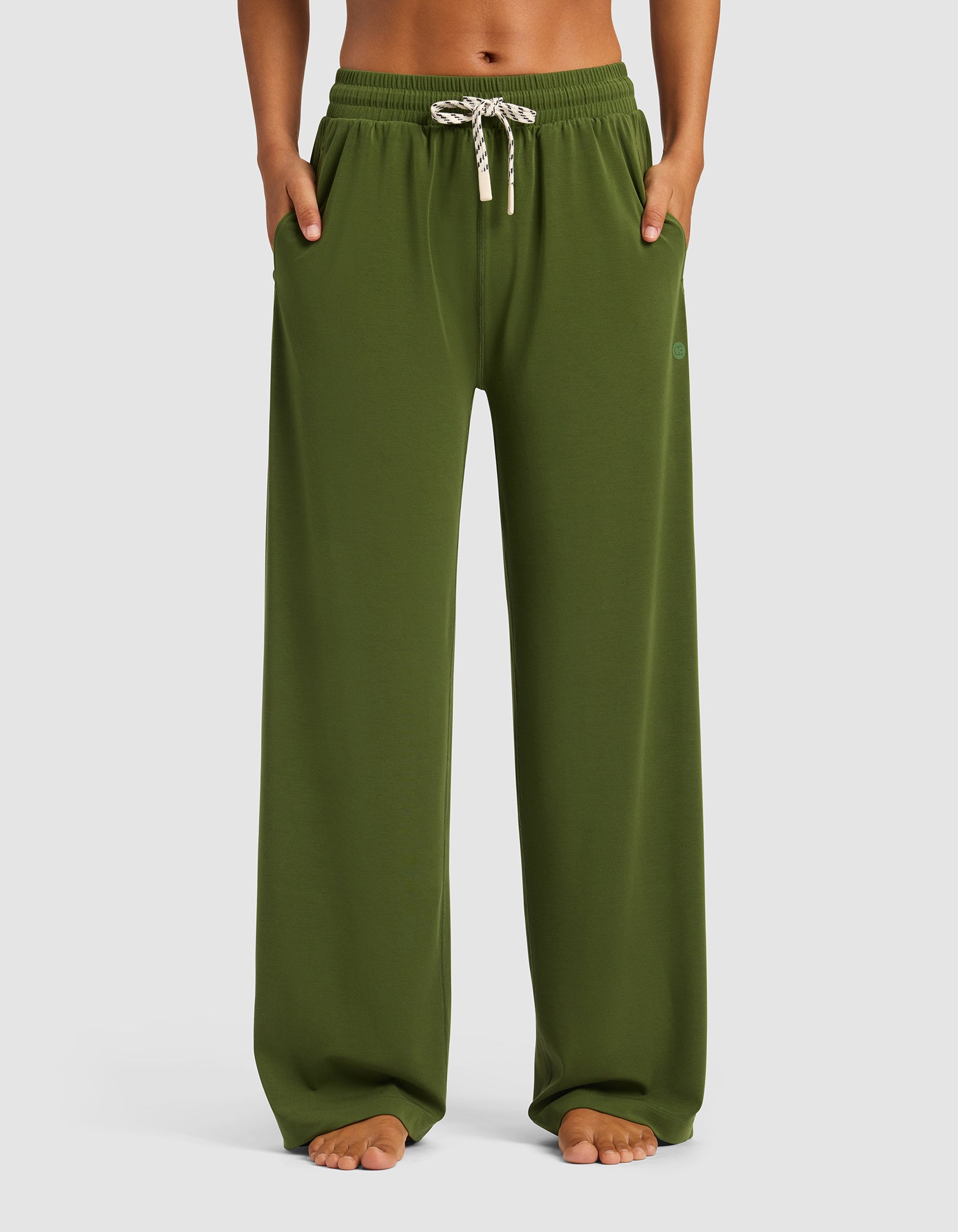 A woman stands barefoot with hands in pockets, wearing Cozy Earth's Women's Studio Wide Leg Pant in olive green, featuring an elastic waistband and white drawstring, against a simple background.