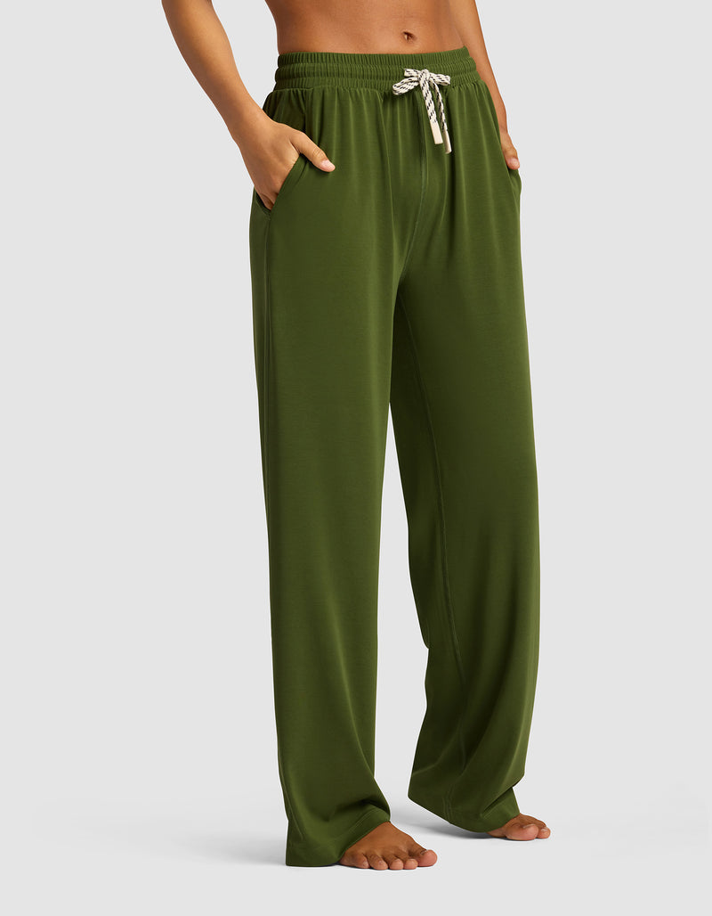 A woman models the Cozy Earth Women's Studio Wide Leg Pant in olive green, featuring a white and gray striped drawstring, hands in pockets, barefoot against a light gray background.