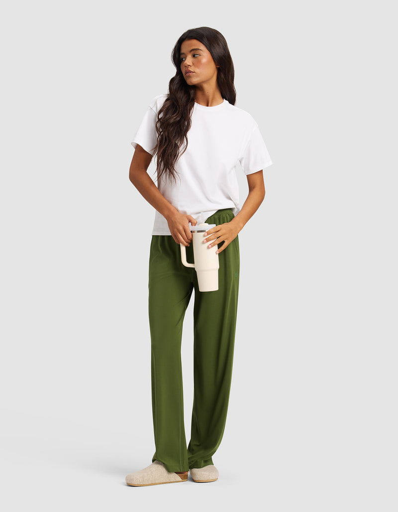 A woman with long brown hair wears a white t-shirt, Cozy Earth Women's Studio Wide Leg Pant in green, and beige slippers, holding a large cream-colored mug against a plain light gray background.