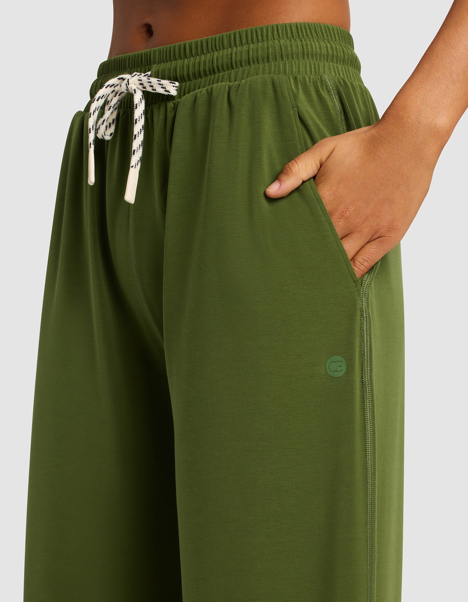 A person wears Cozy Earth Women's Studio Wide Leg Pant in olive green with a white and black drawstring, right hand in the pocket. The image is cropped to show only the waist and upper thigh area.