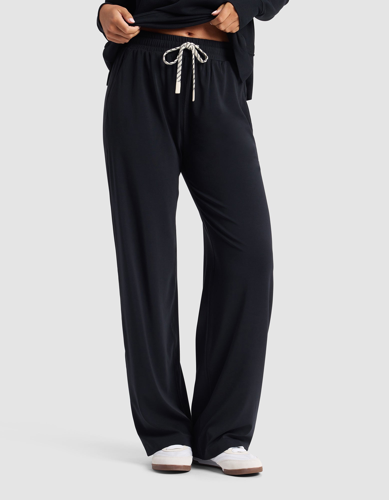A woman wears Cozy Earth's Women's Studio Wide Leg Pant in black with a white drawstring, paired with a black top and white shoes, standing against a plain light background.|Color:Jet Black