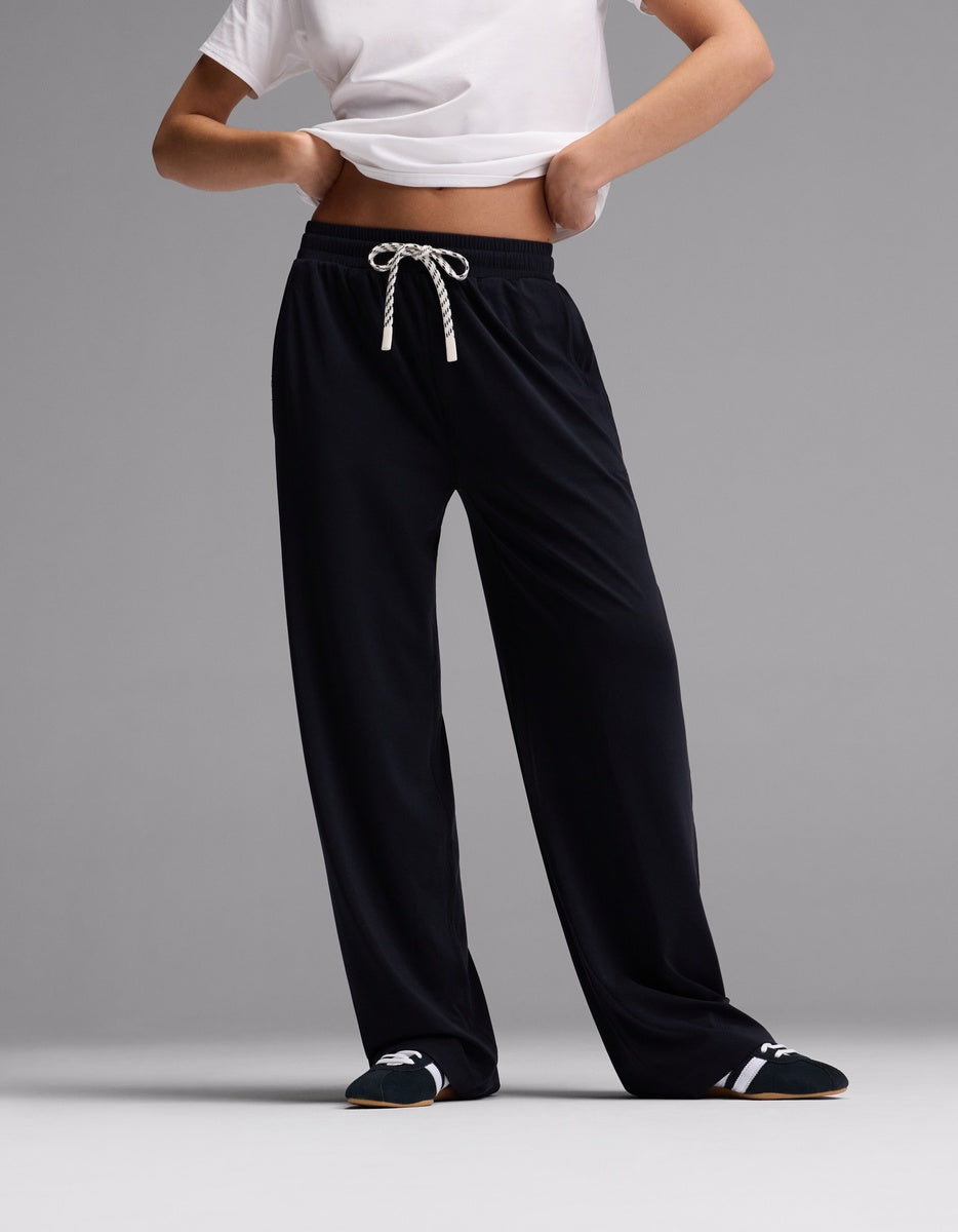 A person stands against a plain gray background wearing Cozy Earth’s Women’s Studio Wide Leg Pant in Jet Black, paired with a cropped white T-shirt and black slippers with white details. Their head and most of their arms are out of frame.|Color:Jet Black