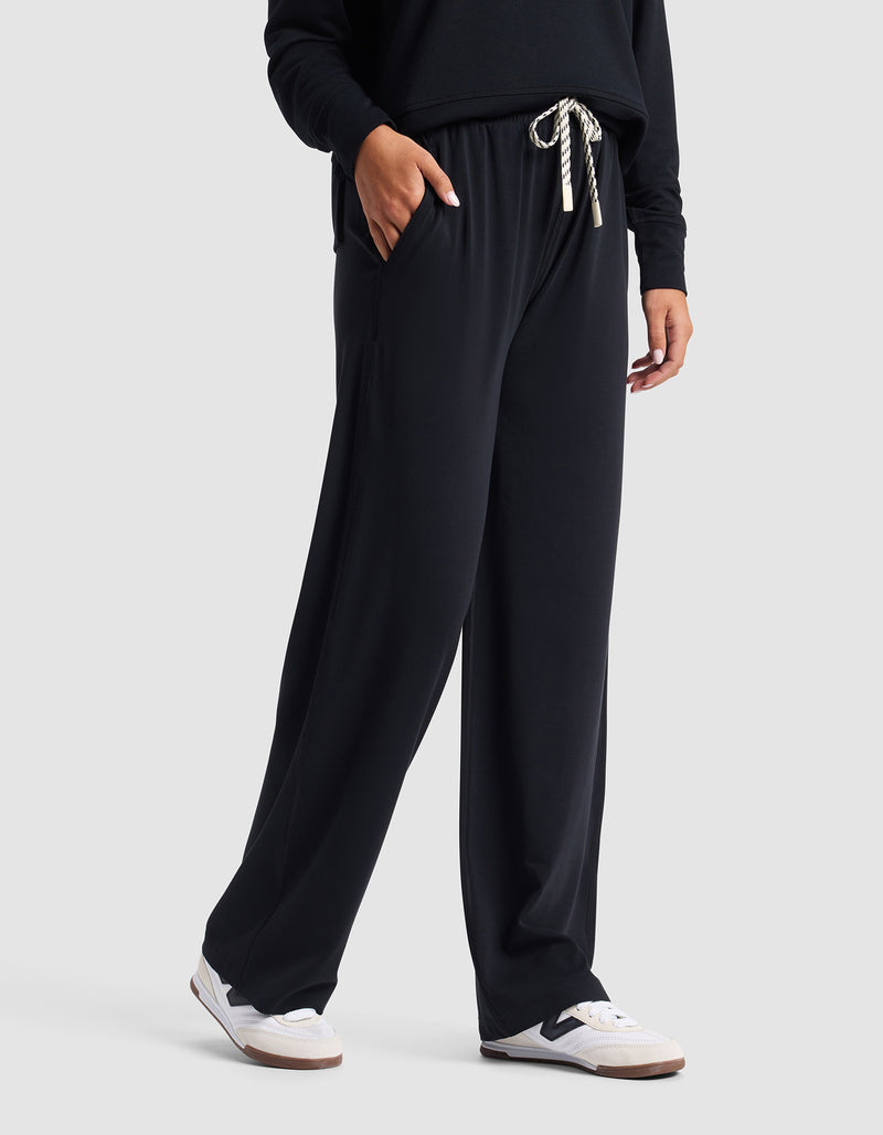 A woman in Cozy Earth's Women's Studio Wide Leg Pant—black with white drawstrings—paired with a matching top and white sneakers, stands with one hand in her pocket against a plain light background.