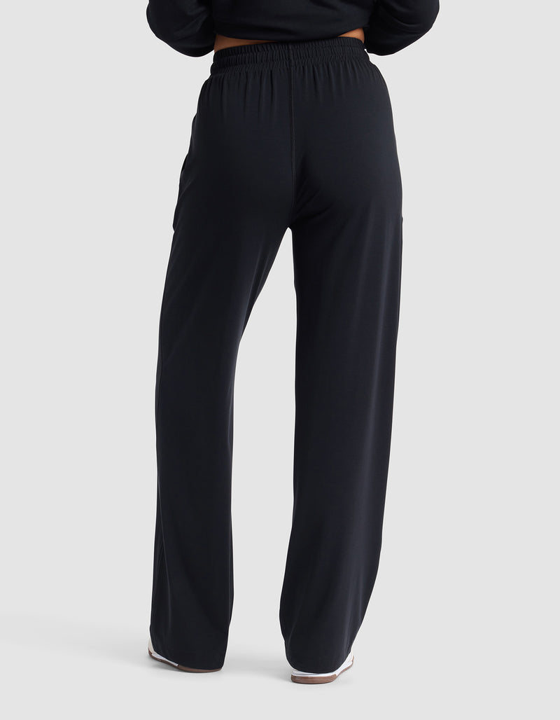 A person stands facing away against a plain light background, showcasing the Cozy Earth Women's Studio Wide Leg Pant in black from the back view.