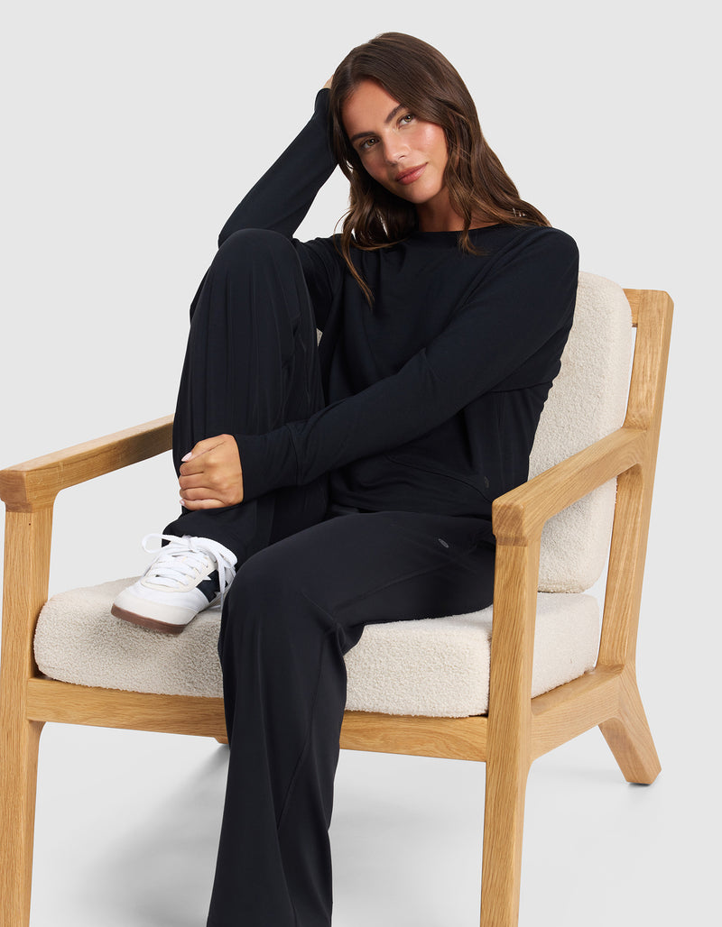 A woman with long brown hair sits sideways on a beige cushioned wooden chair, resting her head on her hand and one knee up. She wears Cozy Earth's Women's Studio Wide Leg Pant, a black long-sleeve top, and white sneakers, smiling softly at the camera.