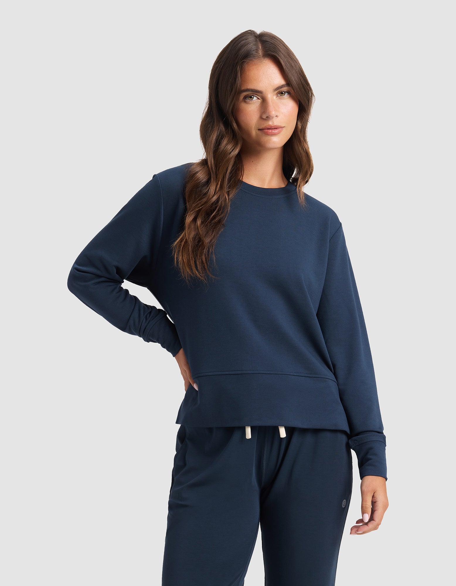 A woman with long brown hair wears the Cozy Earth Women's StudioLite Crewneck and matching navy sweatpants, standing against a plain light gray background with one hand on her hip.|Color:Eclipse