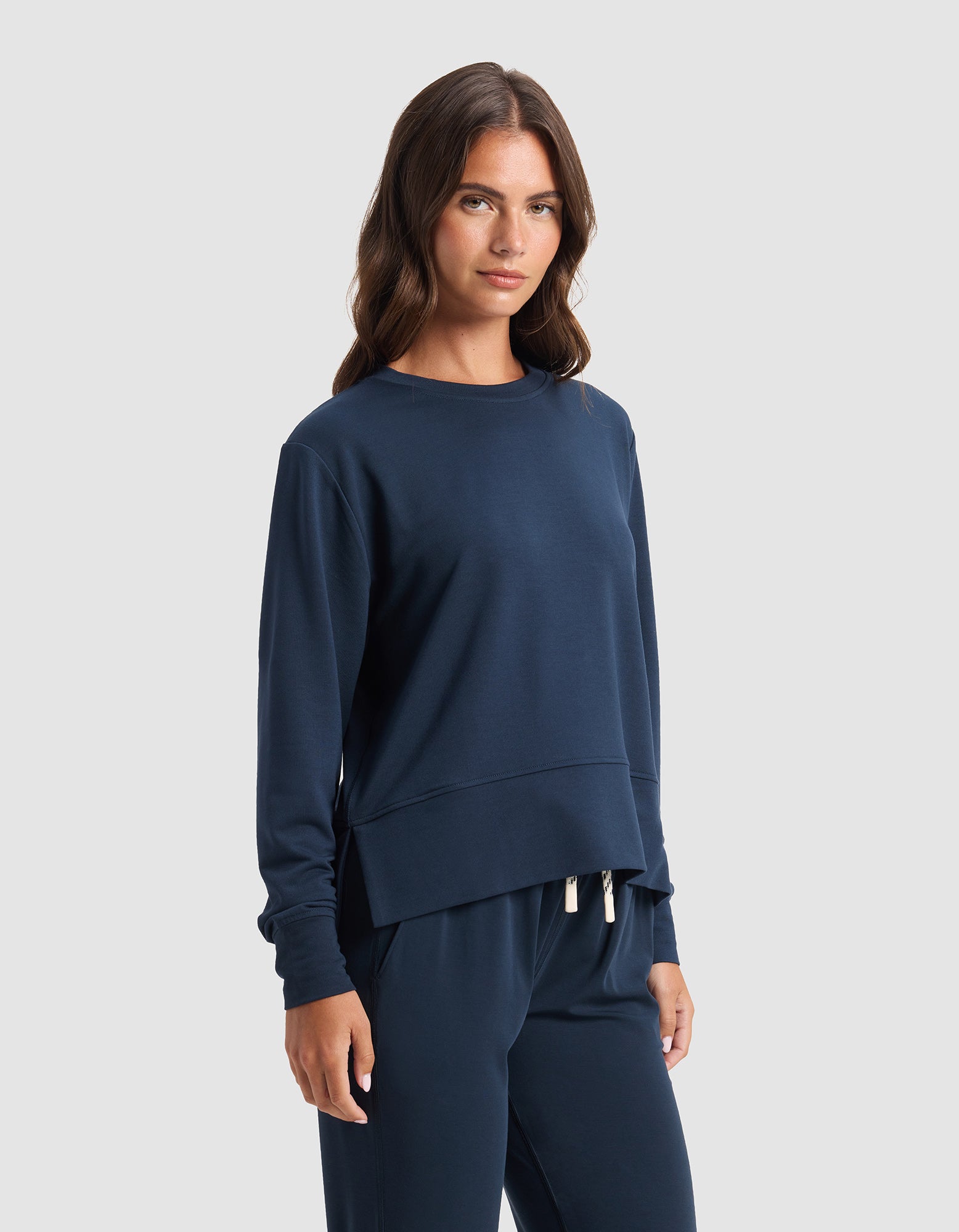 A woman with long brown hair models the Cozy Earth Women's StudioLite Crewneck and matching pants in navy blue, standing against a plain light gray background and looking at the camera with a neutral expression.