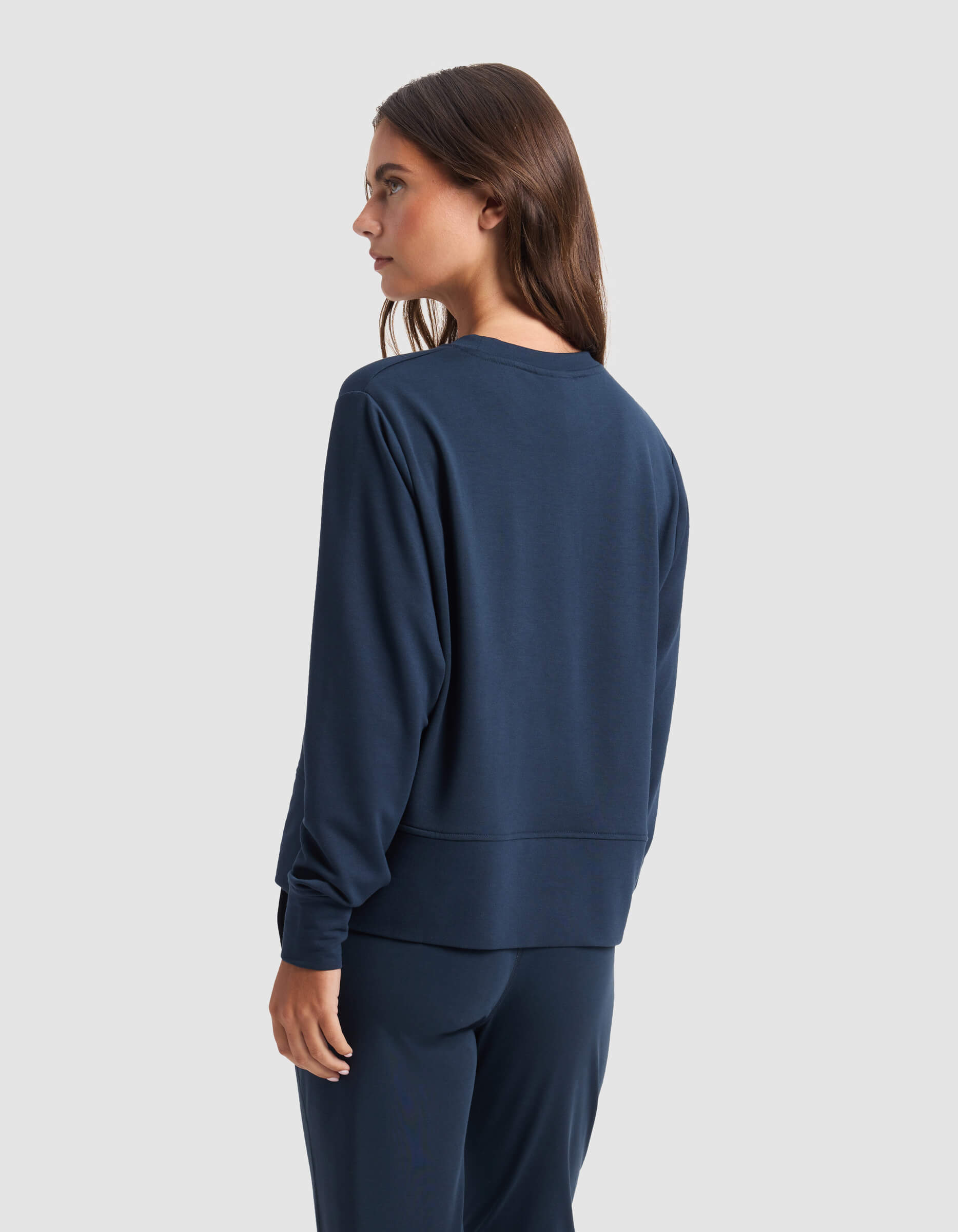 A woman with long brown hair wears the Cozy Earth Women's StudioLite Crewneck and matching pants in navy blue, standing and looking to the side against a plain light gray background.