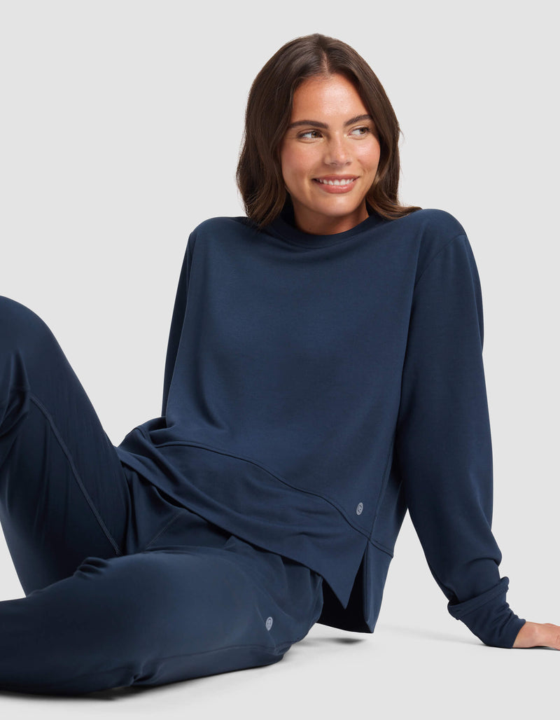 A woman with brown hair, wearing Cozy Earth's Women's StudioLite Crewneck and matching pants in navy blue, sits comfortably on the floor against a light gray background, smiling and looking to the side.