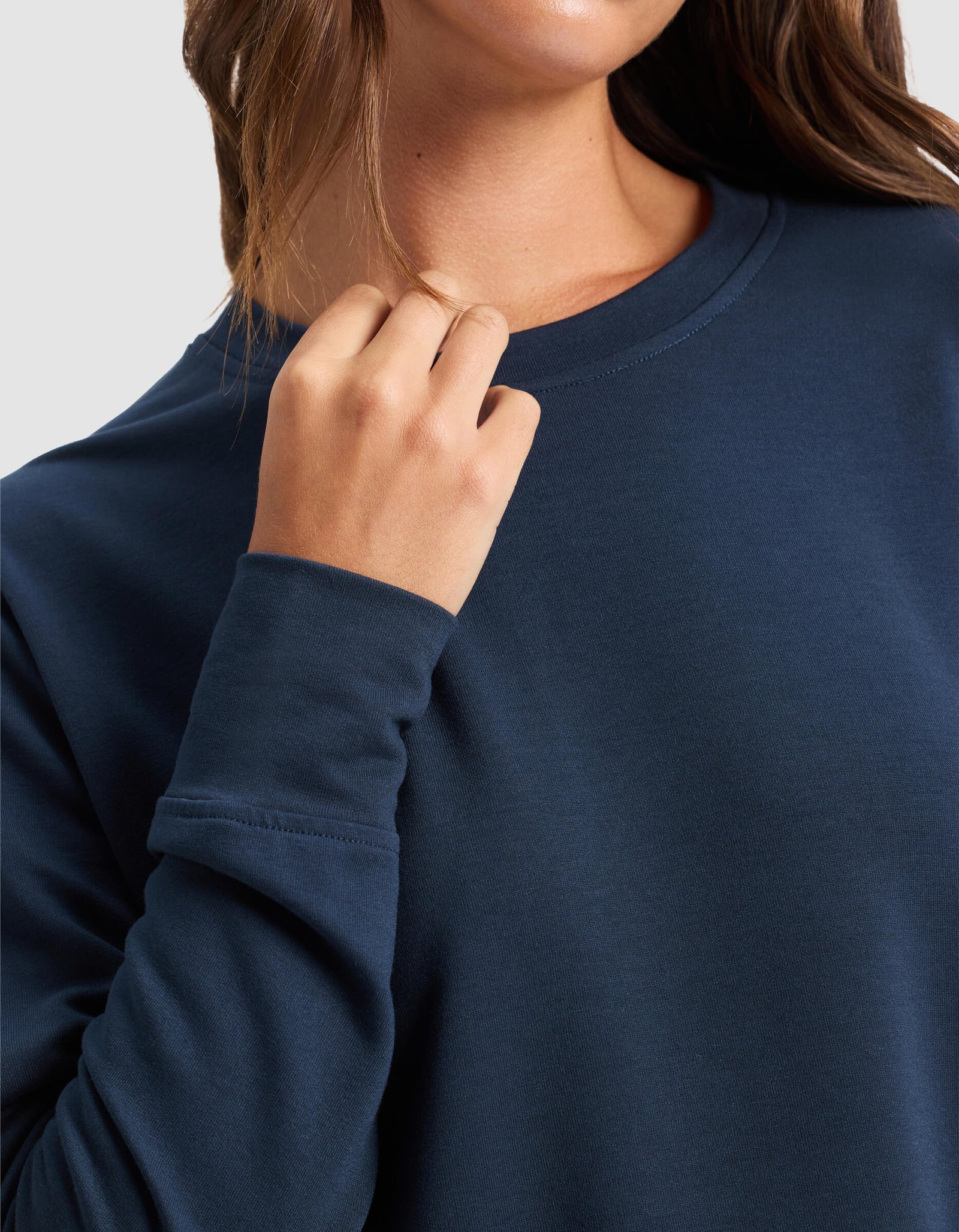 A woman wears the Cozy Earth Women's StudioLite Crewneck, a long-sleeve dark blue shirt. She touches her collar with her right hand, brown hair over her shoulder. Her face is partially out of frame against a plain light background.