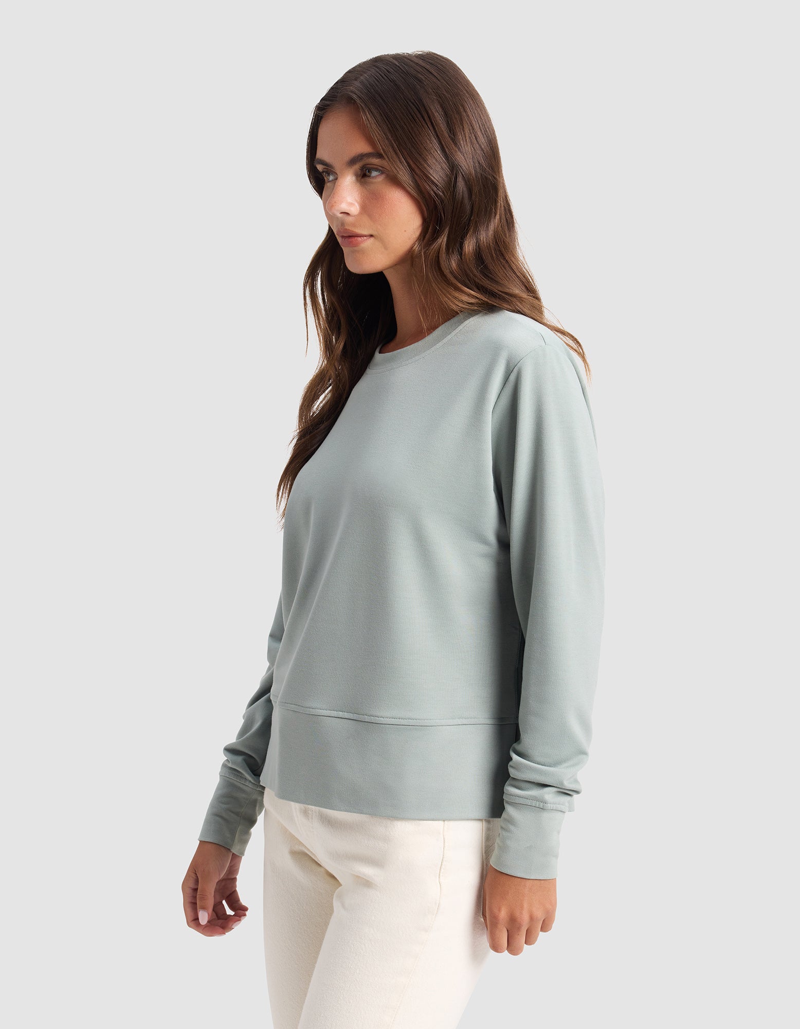 A woman with long brown hair models the Cozy Earth Women's StudioLite Crewneck, a light green long-sleeve top, and off-white pants against a plain light gray background while looking to the side. 