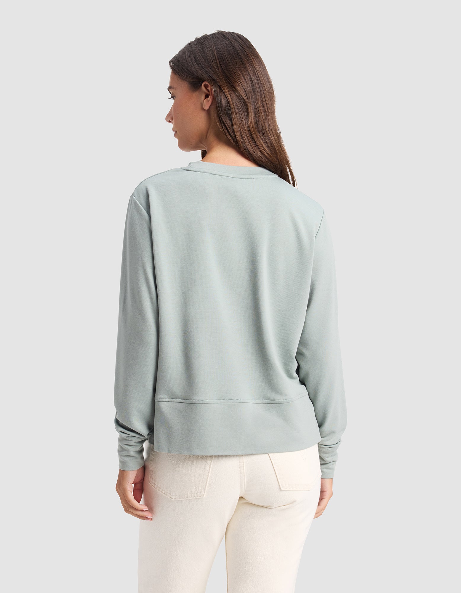 A woman with long brown hair is shown from the back, wearing the Cozy Earth Women's StudioLite Crewneck in light green and off-white pants, standing against a plain light gray background.