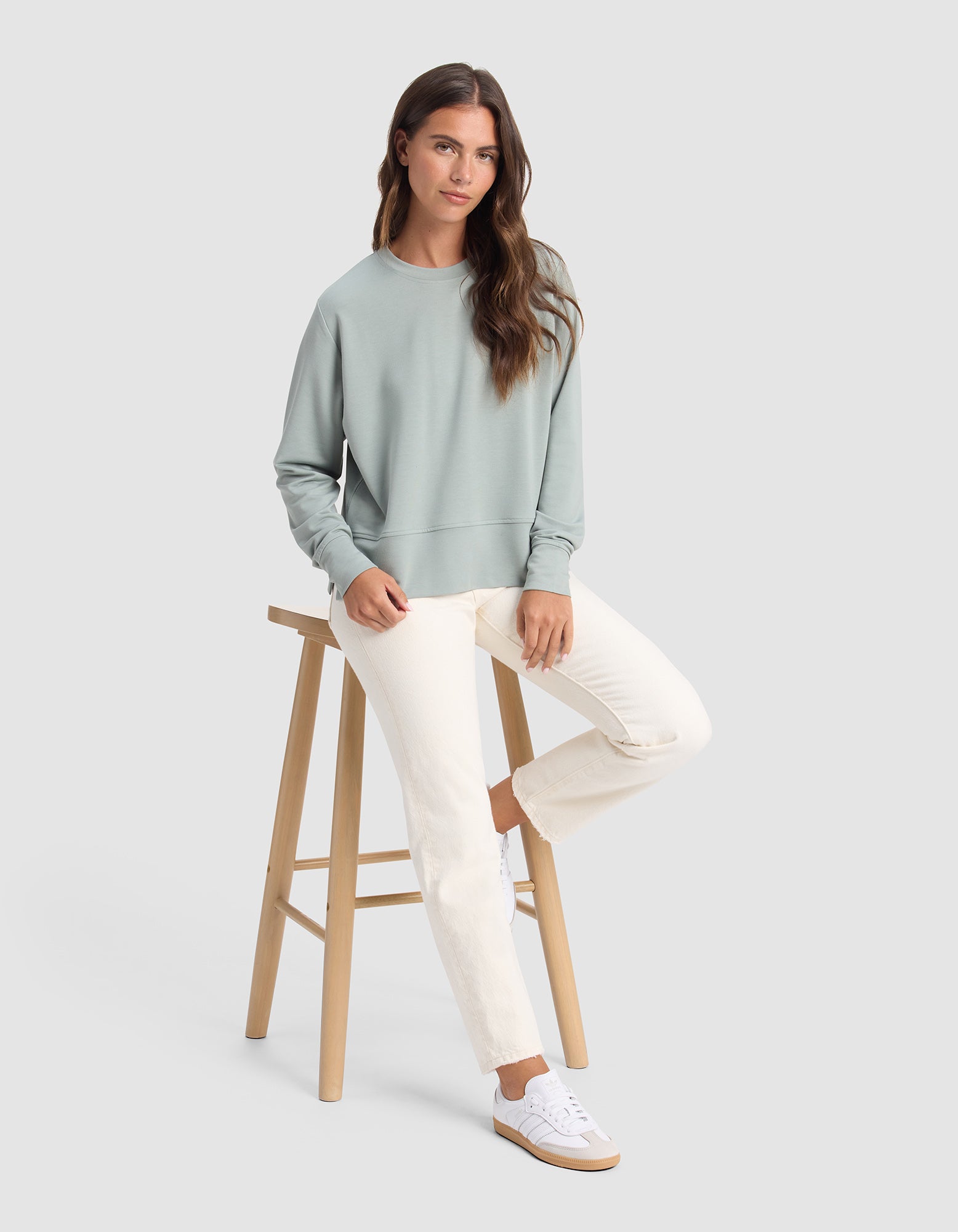 A woman with long brown hair sits on a wooden stool wearing the Cozy Earth Women's StudioLite Crewneck in light green, paired with off-white pants and white sneakers, set against a plain light gray background.