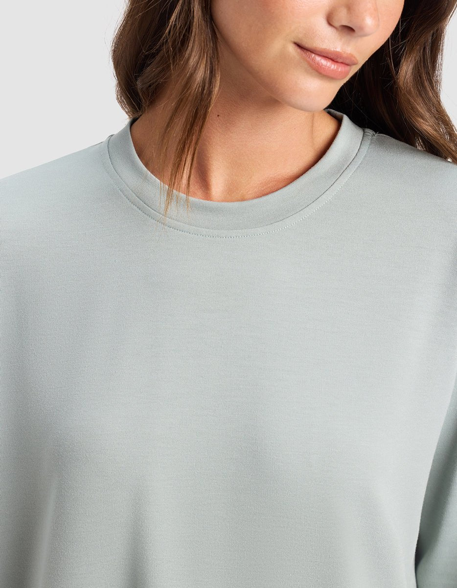 A woman with long brown hair wears the Cozy Earth Women's StudioLite Crewneck in light grey, shown from the shoulders up against a plain background. Her face is partially visible, and she has a slight smile.