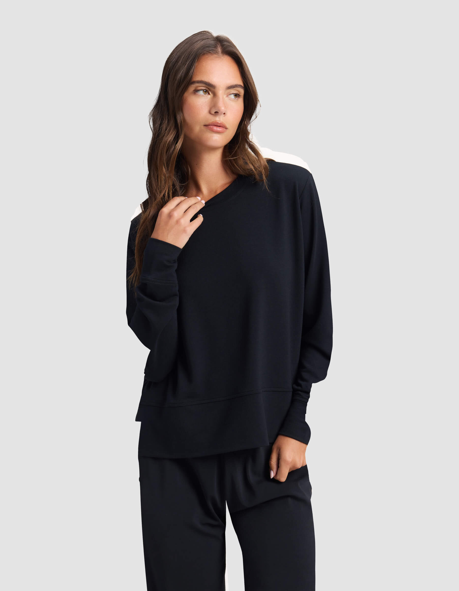 A woman with long brown hair wears the Cozy Earth Women's StudioLite Crewneck, a loose-fitting black sweatshirt with white shoulder accents, and matching black pants, standing against a plain light gray background.