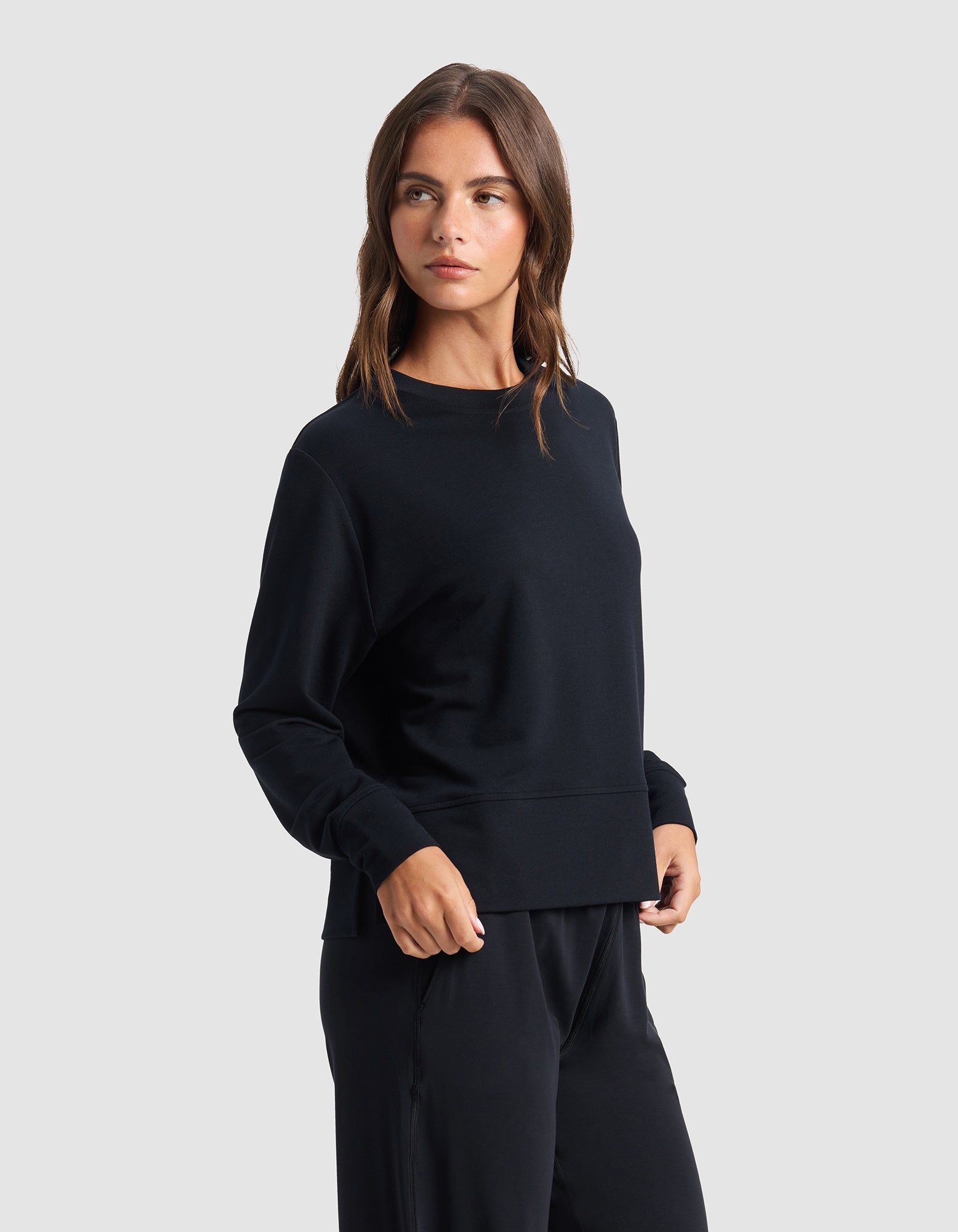 A woman with long brown hair wears the Cozy Earth Women's StudioLite Crewneck in black with matching pants, standing against a plain light gray background and looking slightly to her left.
