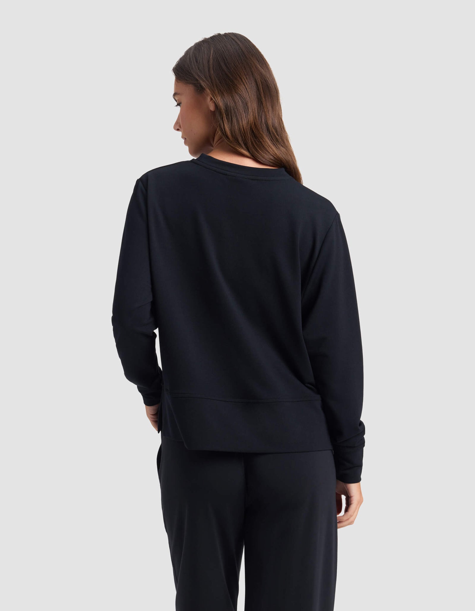 A woman with long brown hair is seen from behind, wearing the Cozy Earth Women's StudioLite Crewneck and black pants, standing against a plain light gray background.