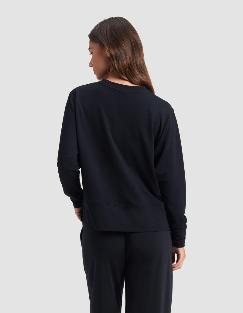 A woman with long brown hair is seen from behind, wearing the Cozy Earth Women's StudioLite Crewneck and black pants, standing against a plain light gray background.