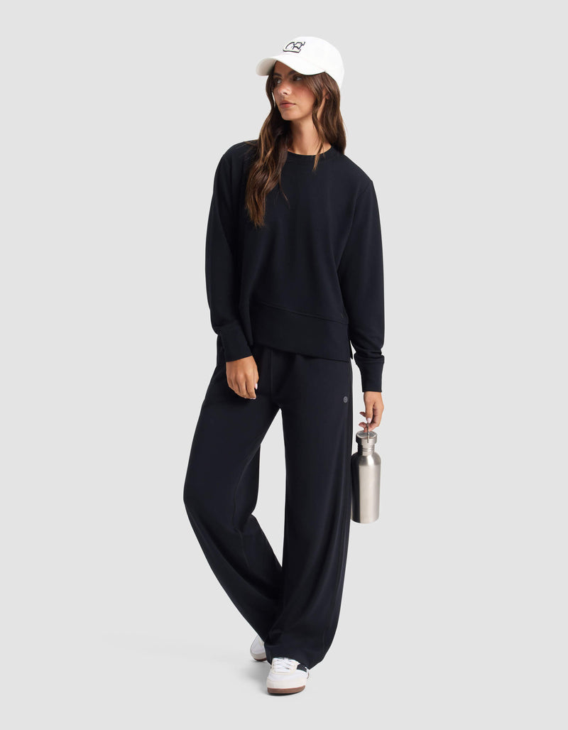 A woman in the Cozy Earth Women's StudioLite Crewneck stands relaxed against a plain background, paired with black wide-leg pants, a white cap, and white sneakers while holding a metal water bottle and looking to the side.