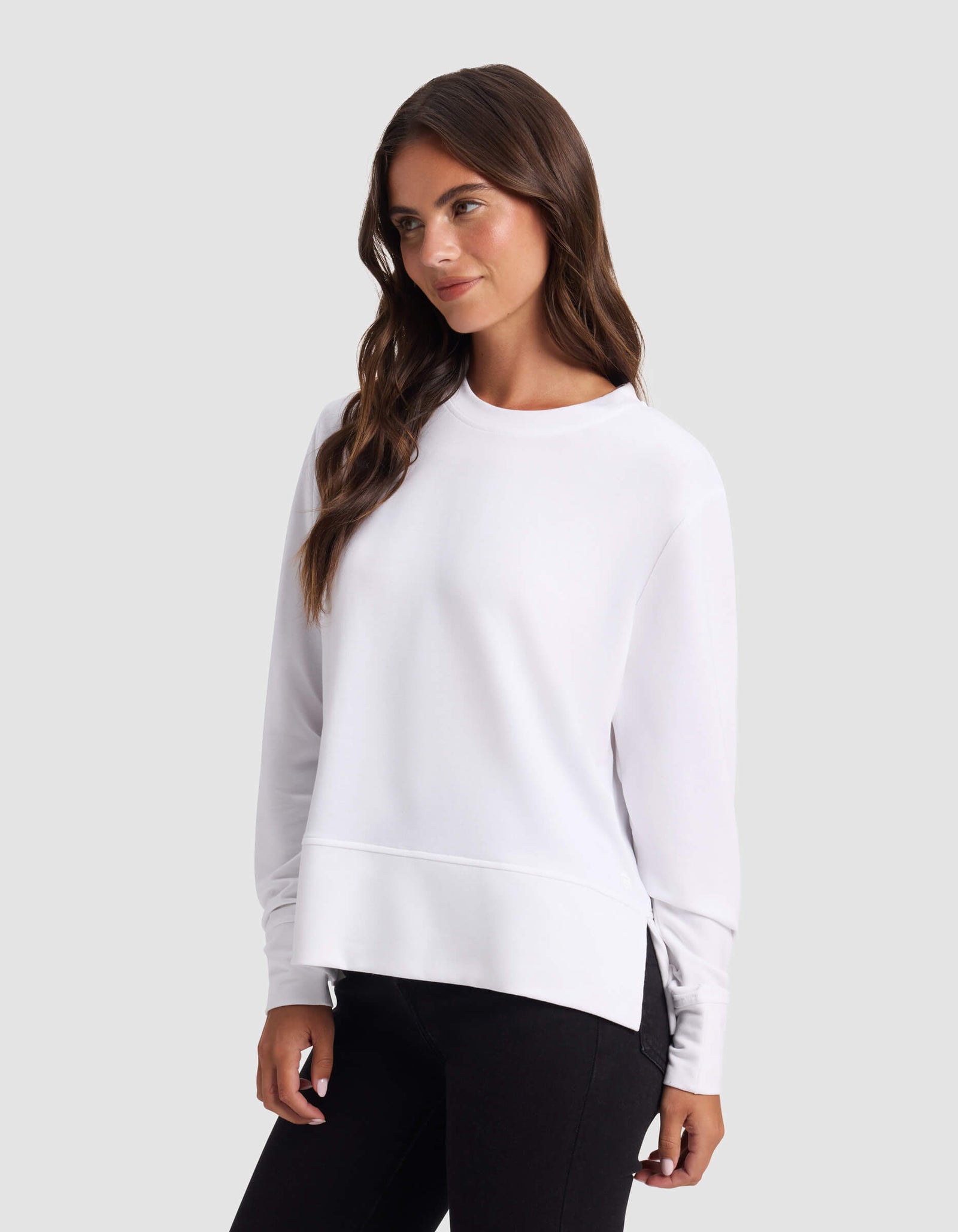 A woman with long brown hair wears the Cozy Earth Women's StudioLite Crewneck in white and black pants, standing against a light gray background and looking slightly to the side with a relaxed expression.