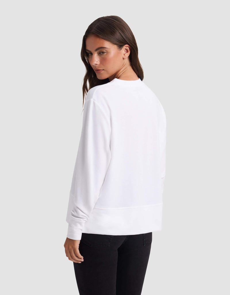 A woman with long brown hair models the Cozy Earth Women's StudioLite Crewneck in white with black pants, standing against a plain light gray background and looking over her left shoulder.