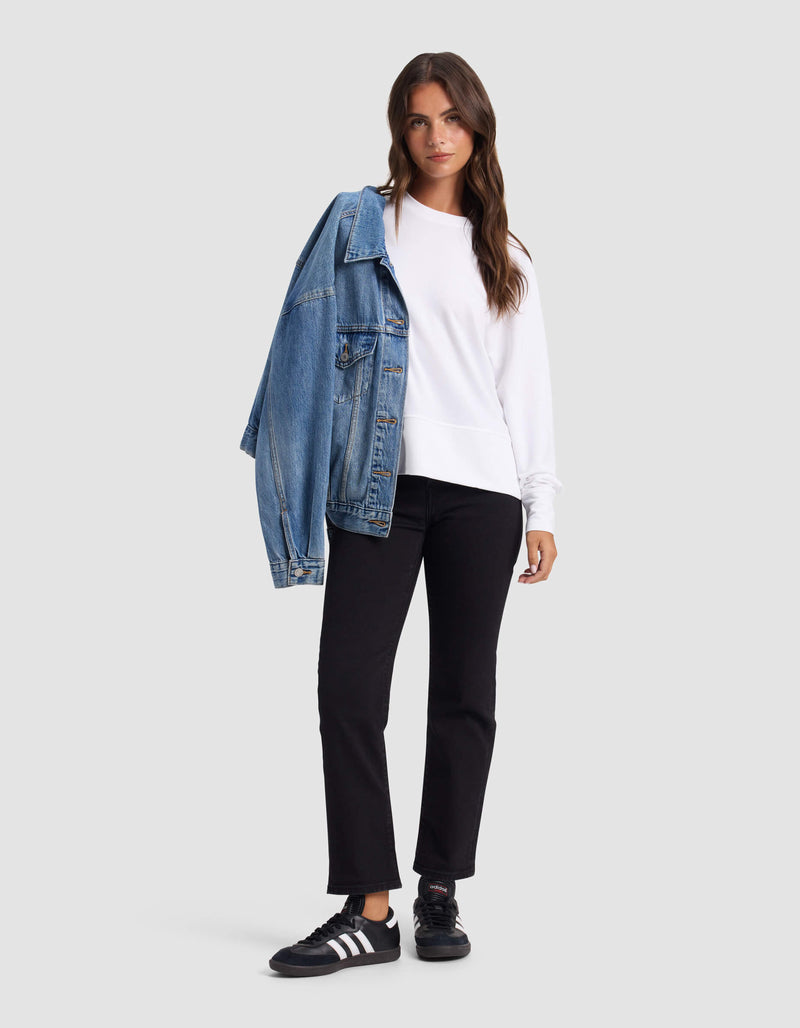 A woman stands against a plain light gray background, wearing a Cozy Earth Women's StudioLite Crewneck, black pants, black Adidas sneakers, and a blue denim jacket draped over one shoulder.