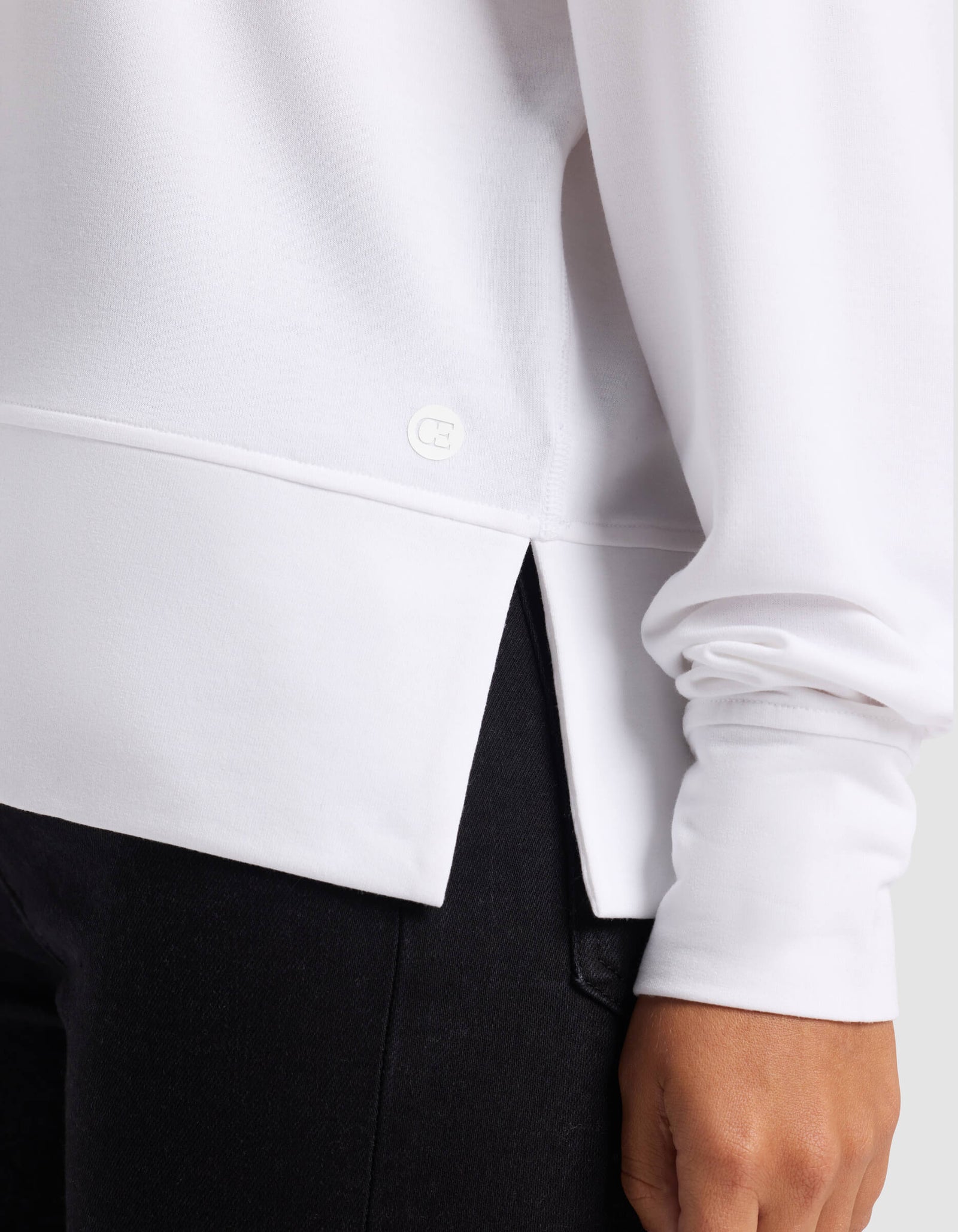 A close-up of a person wearing the Cozy Earth Women's StudioLite Crewneck in white, featuring a side slit at the hem, paired with black pants. The person's hand rests by their side.