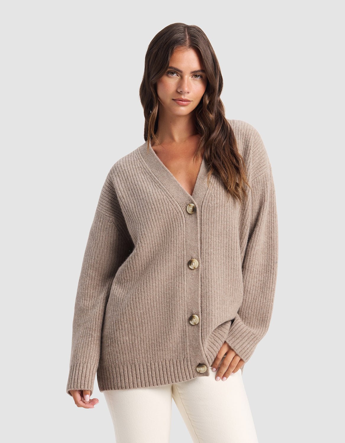 A woman with long brown hair models the Cozy Earth Oversized Sunday Cashmere Cardigan in beige, featuring large buttons, over cream pants against a plain gray background.