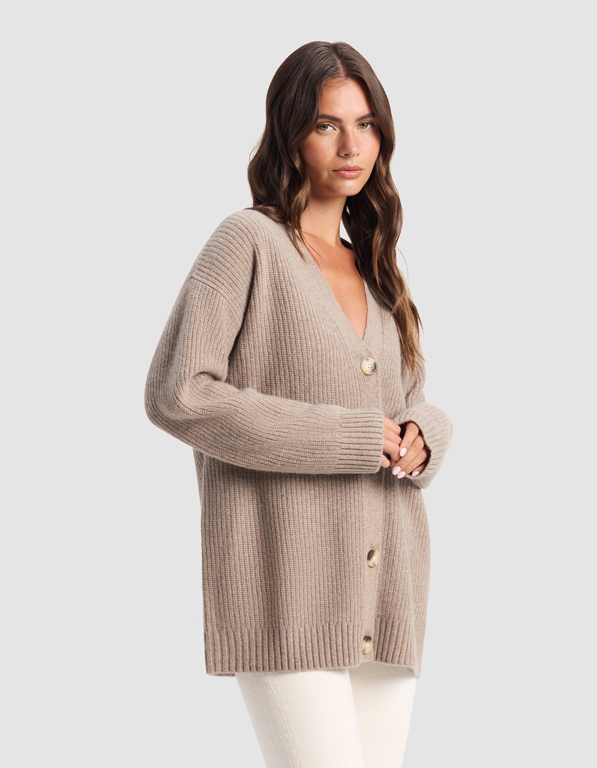 A woman with long brown hair wears Cozy Earth's Oversized Sunday Cashmere Cardigan in beige with large buttons and white pants, standing against a light gray background.