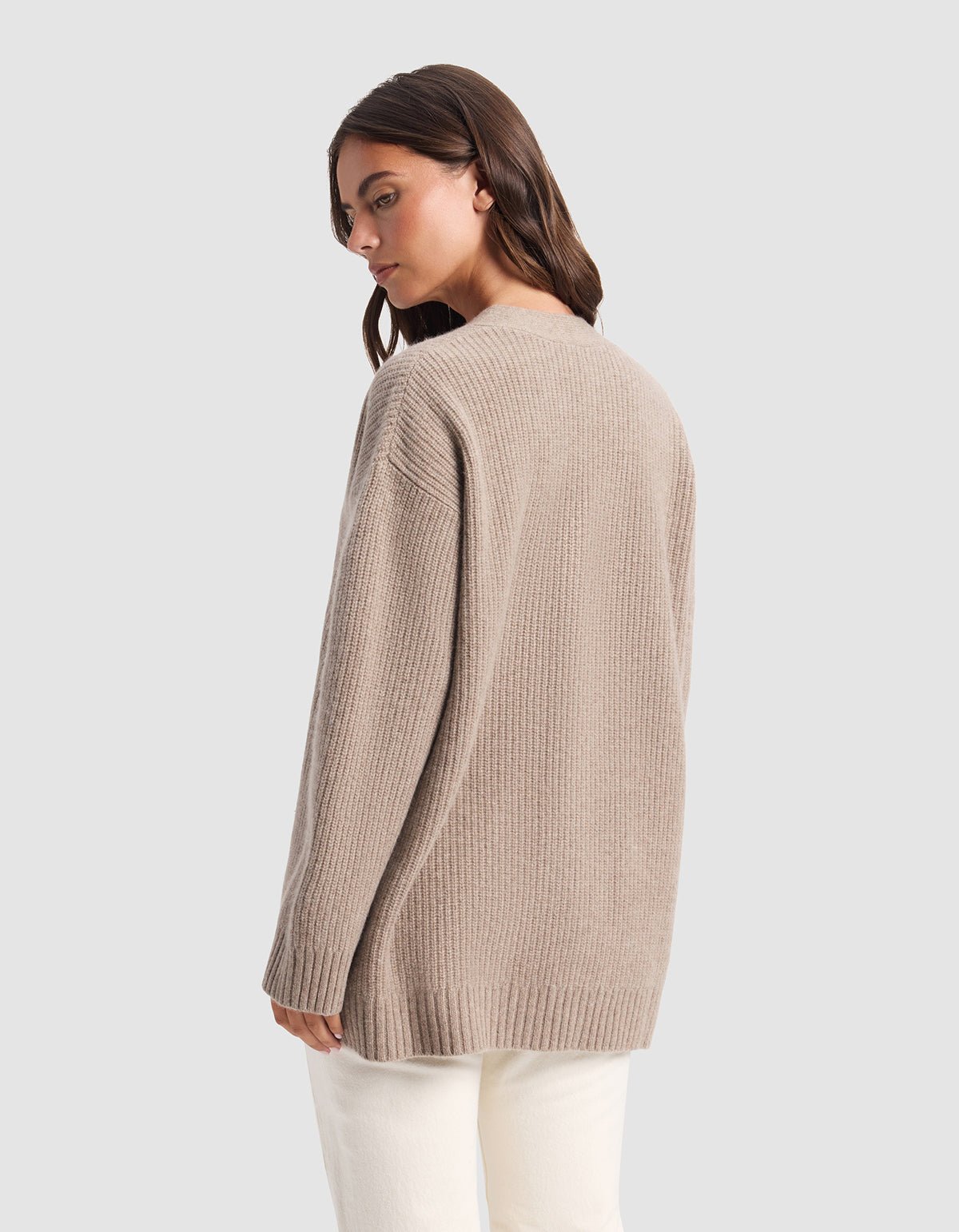 A woman with long brown hair stands facing away, head turned left, wearing Cozy Earth’s Oversized Sunday Cashmere Cardigan in light brown and white pants against a plain gray background.
