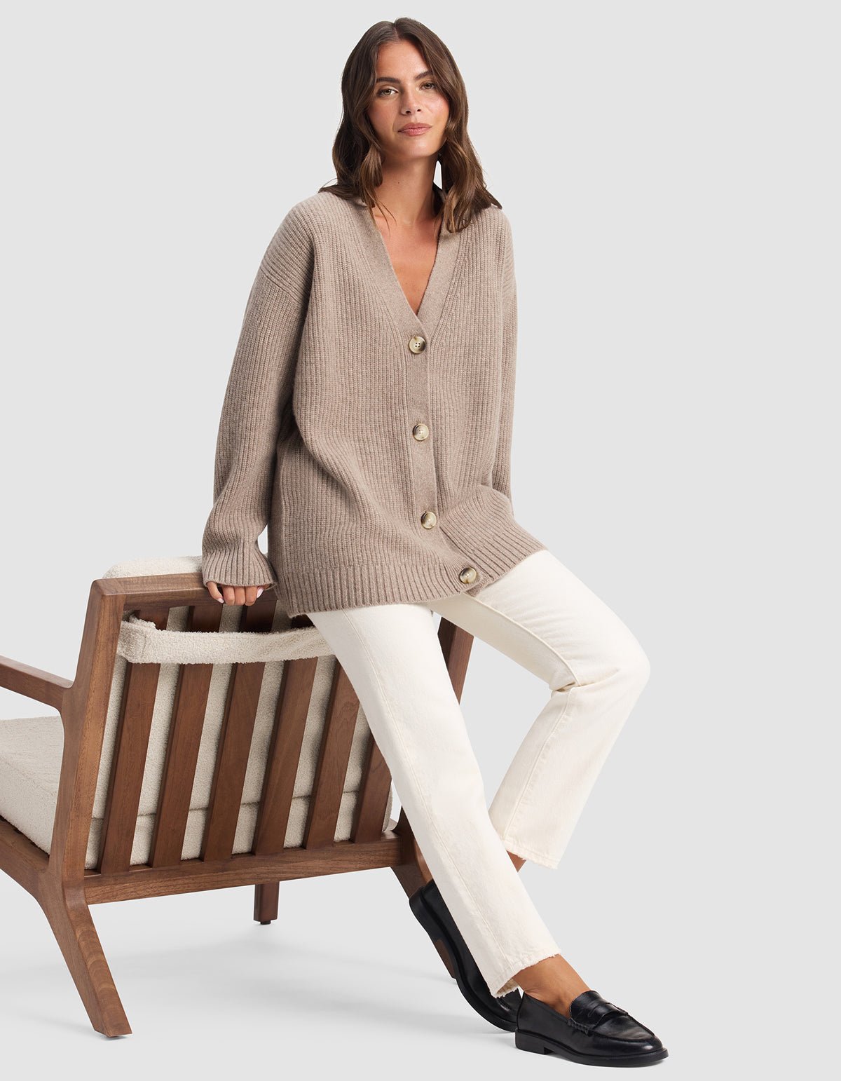A woman with wavy brown hair sits on a wooden chair, wearing Cozy Earth's Oversized Sunday Cashmere Cardigan in beige with large buttons, paired with white pants and black loafers, against a plain light gray background.