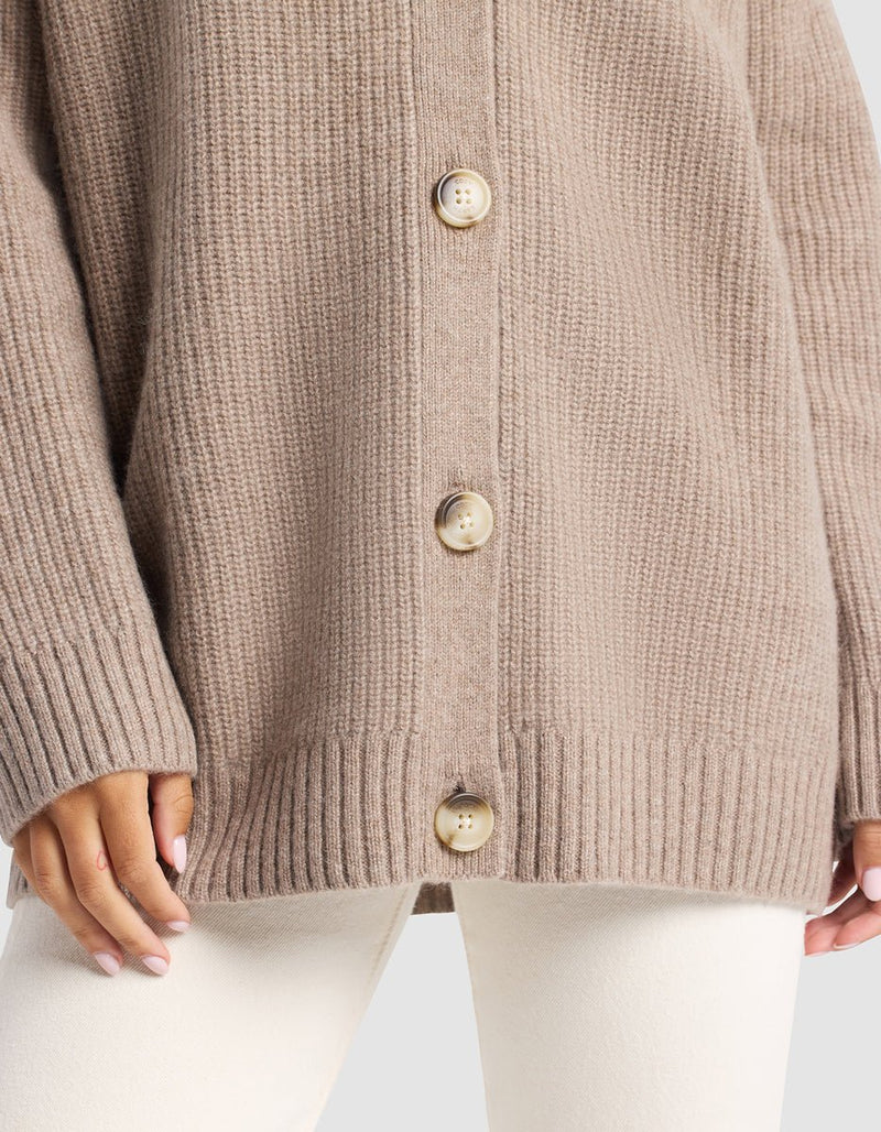 A close-up shows the lower part of the Cozy Earth Oversized Sunday Cashmere Cardigan in beige, featuring ribbed knit and large buttons, paired with cream pants. Only the hands and cardigan are visible.